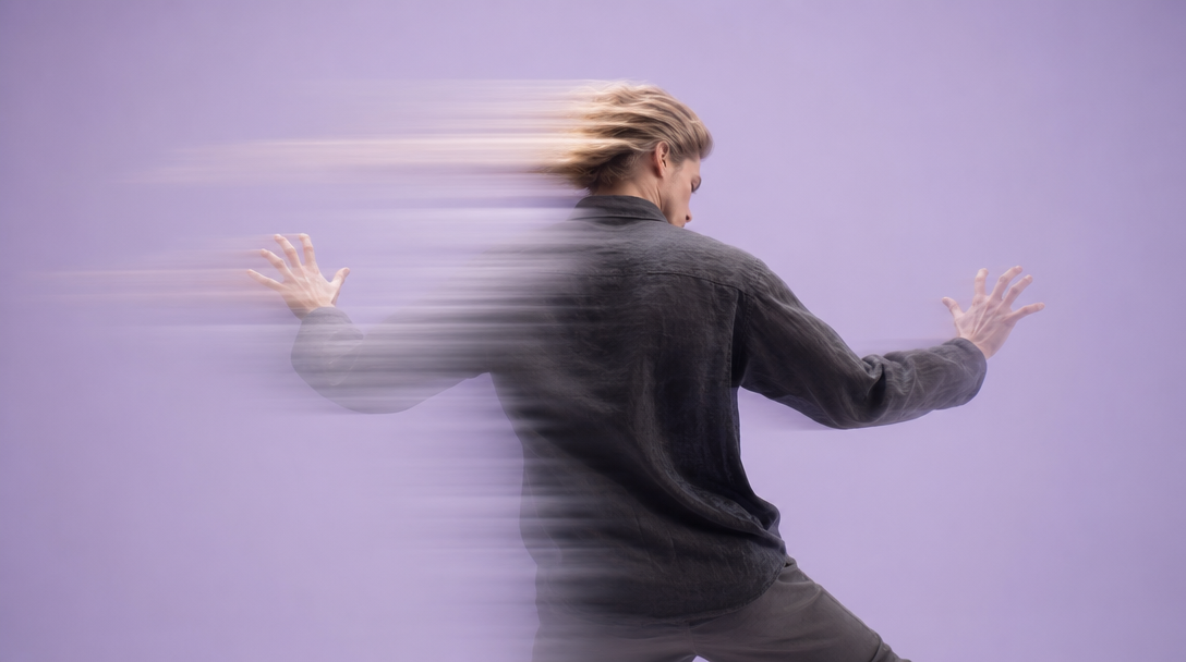 A person mid-step in a lilac studio, arms slightly raised with fingers spread wide in a gesture of examination, viewed from behind at slight angle,