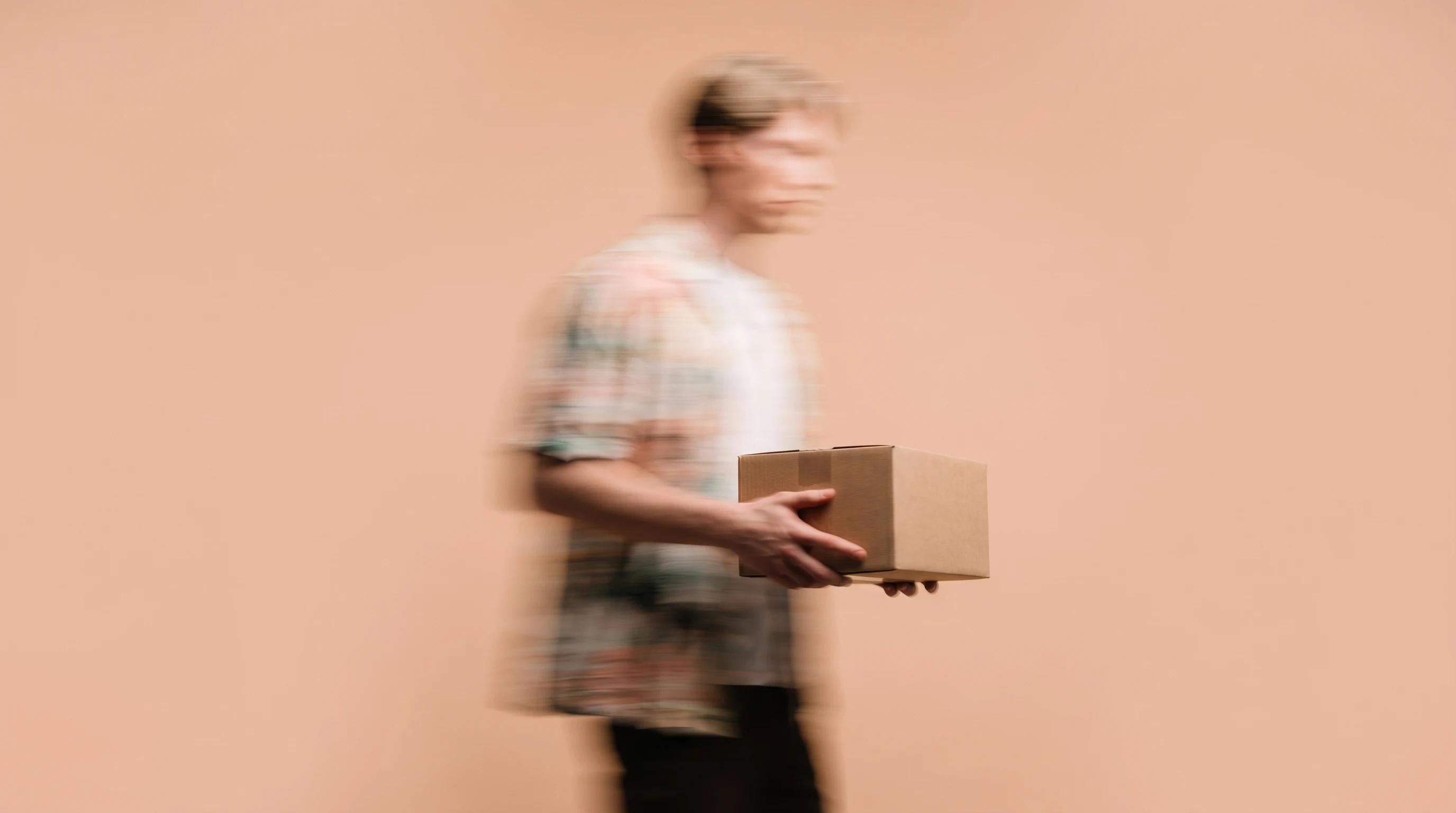 Person mid-pivot in a peach-backdrop studio, three-quarter framing, holding a small unbranded product box at waist height