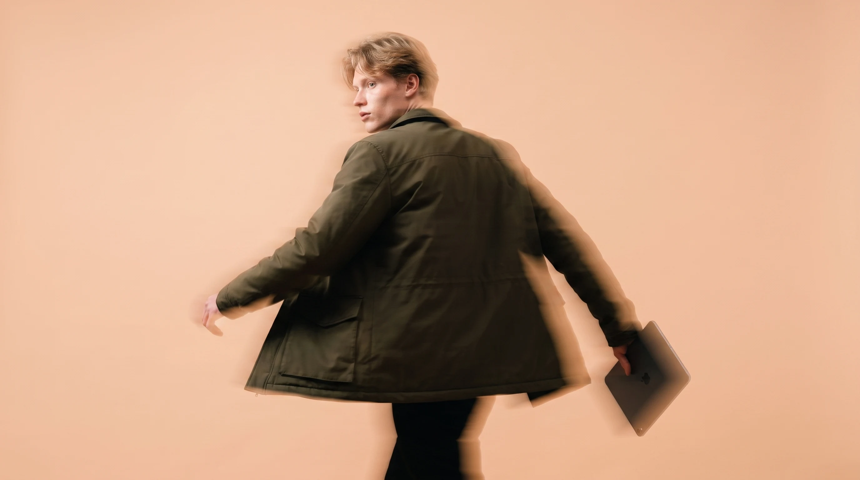 A person mid-step through a peach-backdrop studio, holding a slim tablet loosely at their side, glancing back over one shoulder, wearing a dark olive