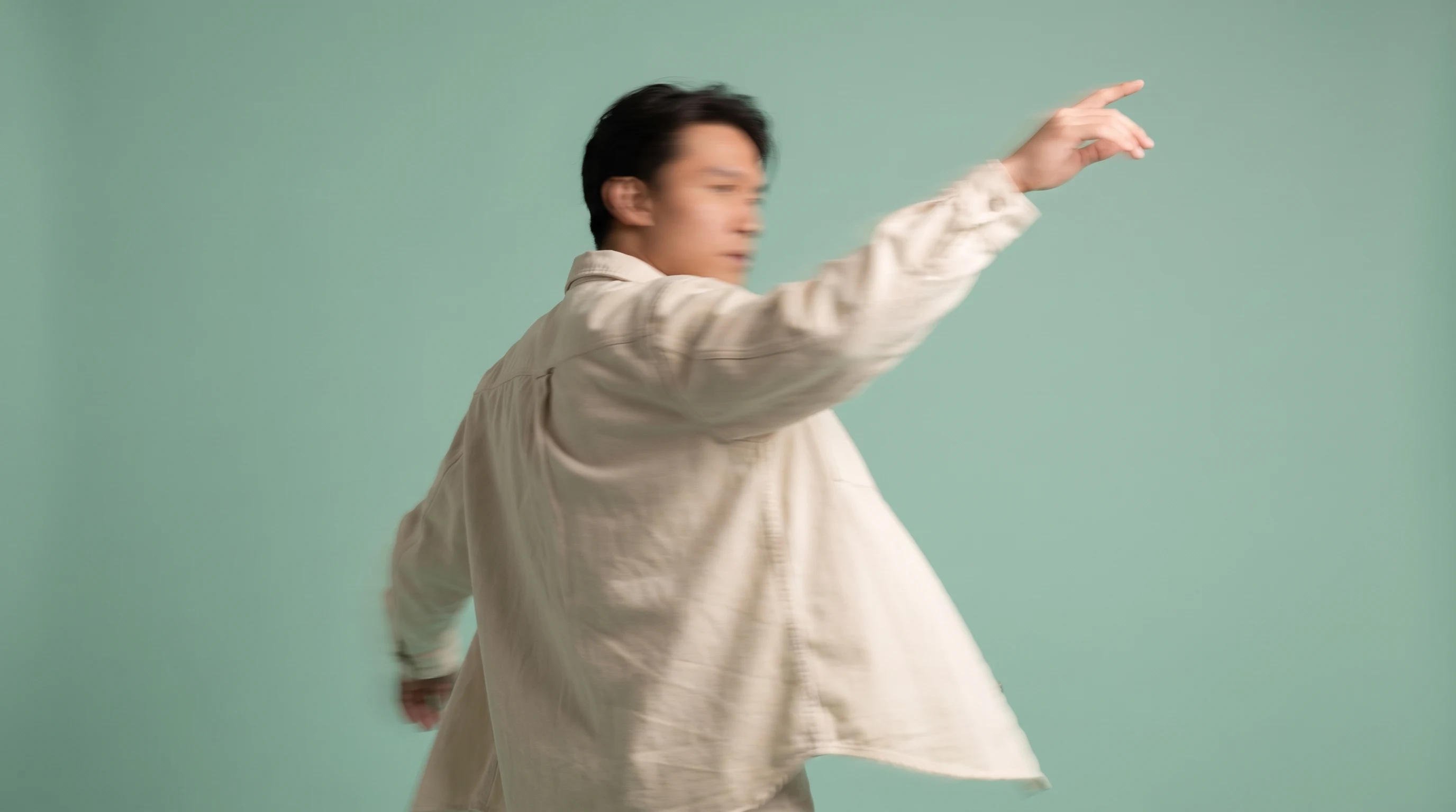 A person mid-turn in a mint-backdrop studio, one arm raised gesturing toward an unseen screen, wearing a loose cream overshirt, three-quarter framing