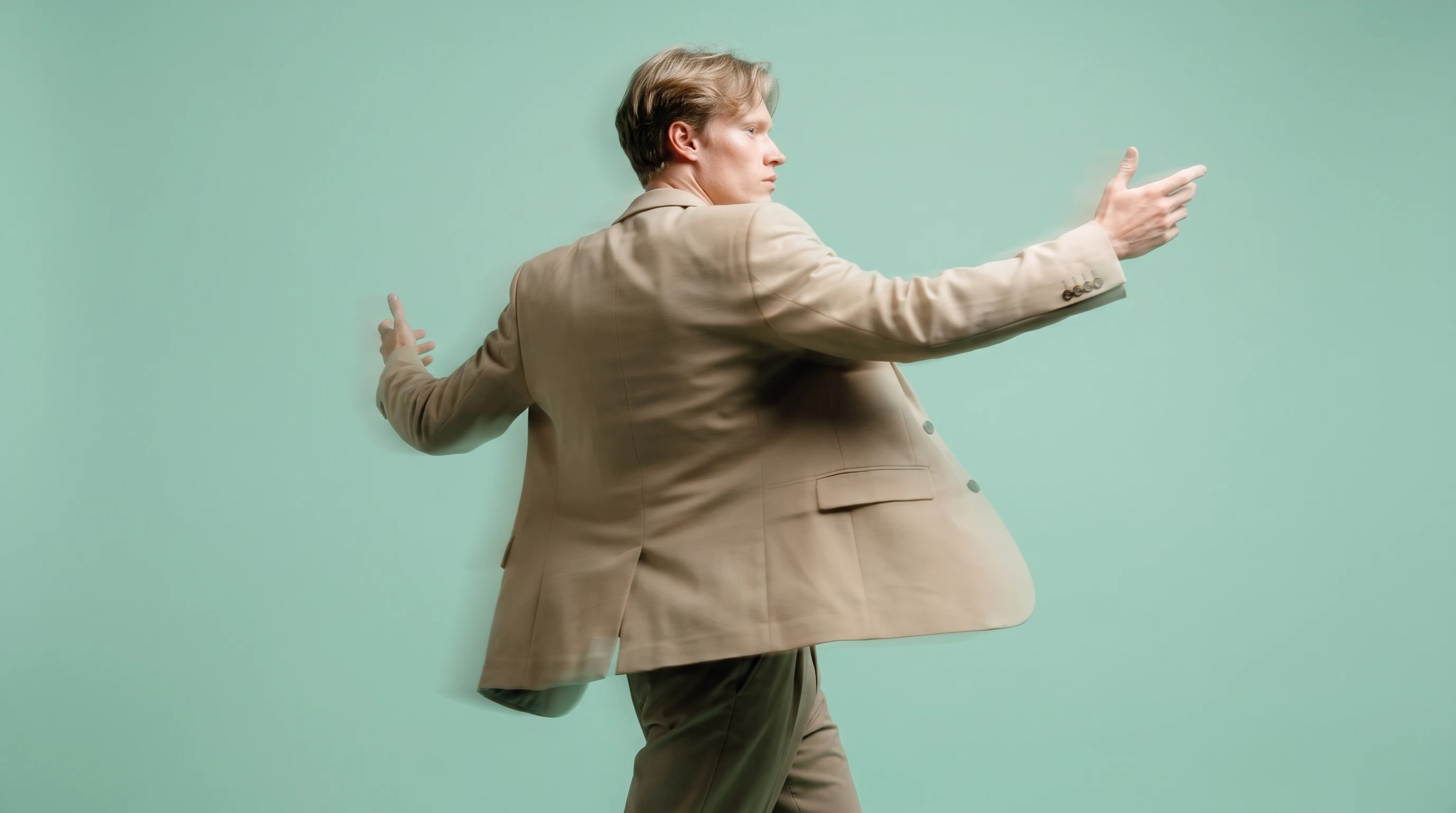 A person mid-turn in a mint-backdrop studio, arms gesturing outward as if presenting an unseen mood board, wearing a tailored sand-coloured blazer,