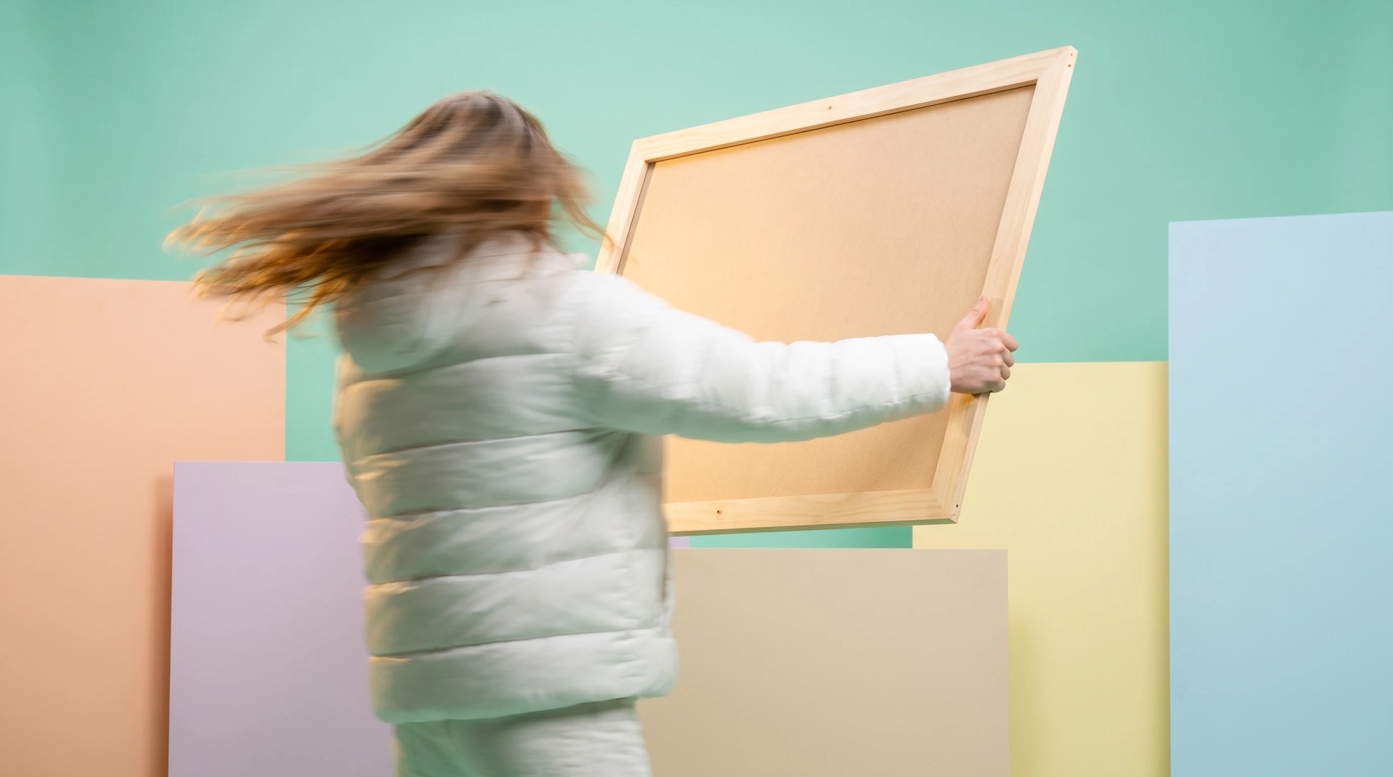 a person mid-turn in a mint-green studio, holding a large unbranded storyboard frame up to the light, coat mid-sweep behind them