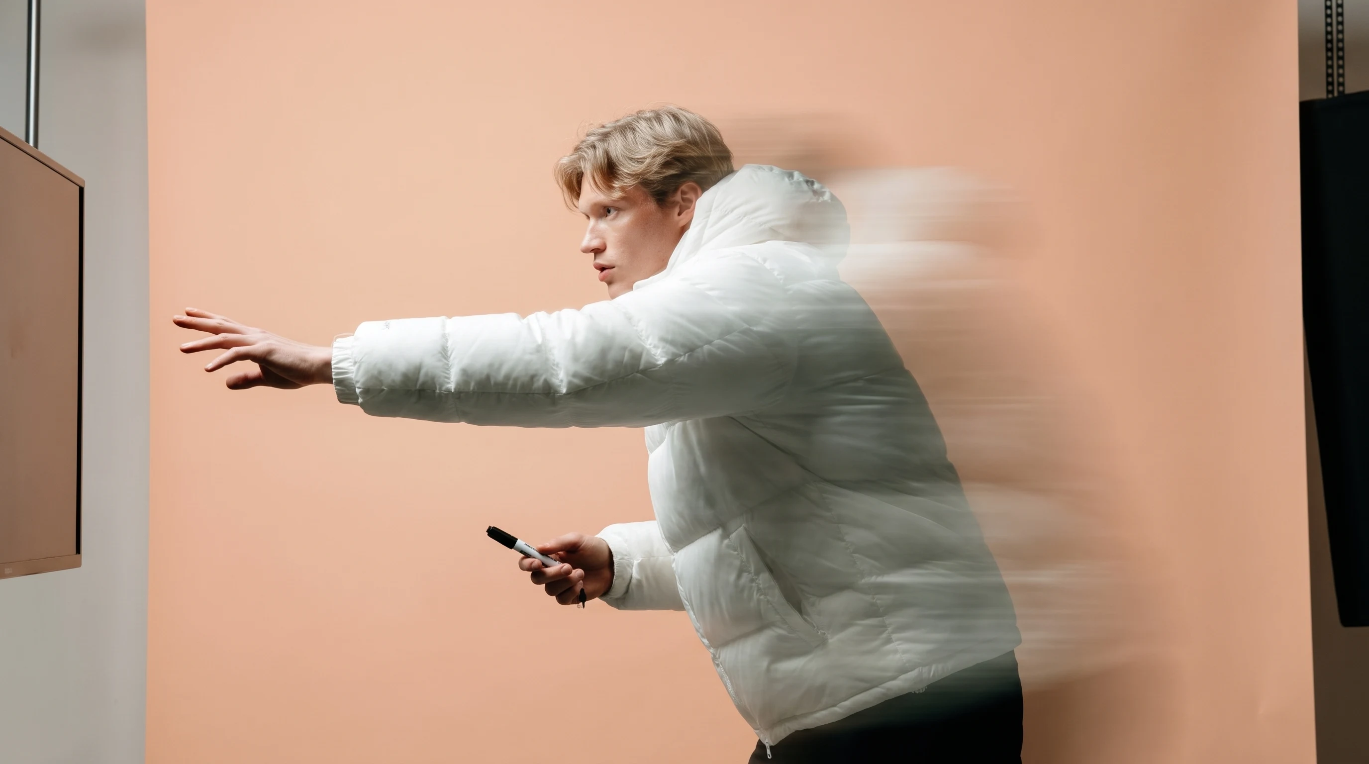 A person mid-reach in a peach-backdrop studio gesturing toward an unseen screen with one arm extended, other hand gripping a marker, slight forward