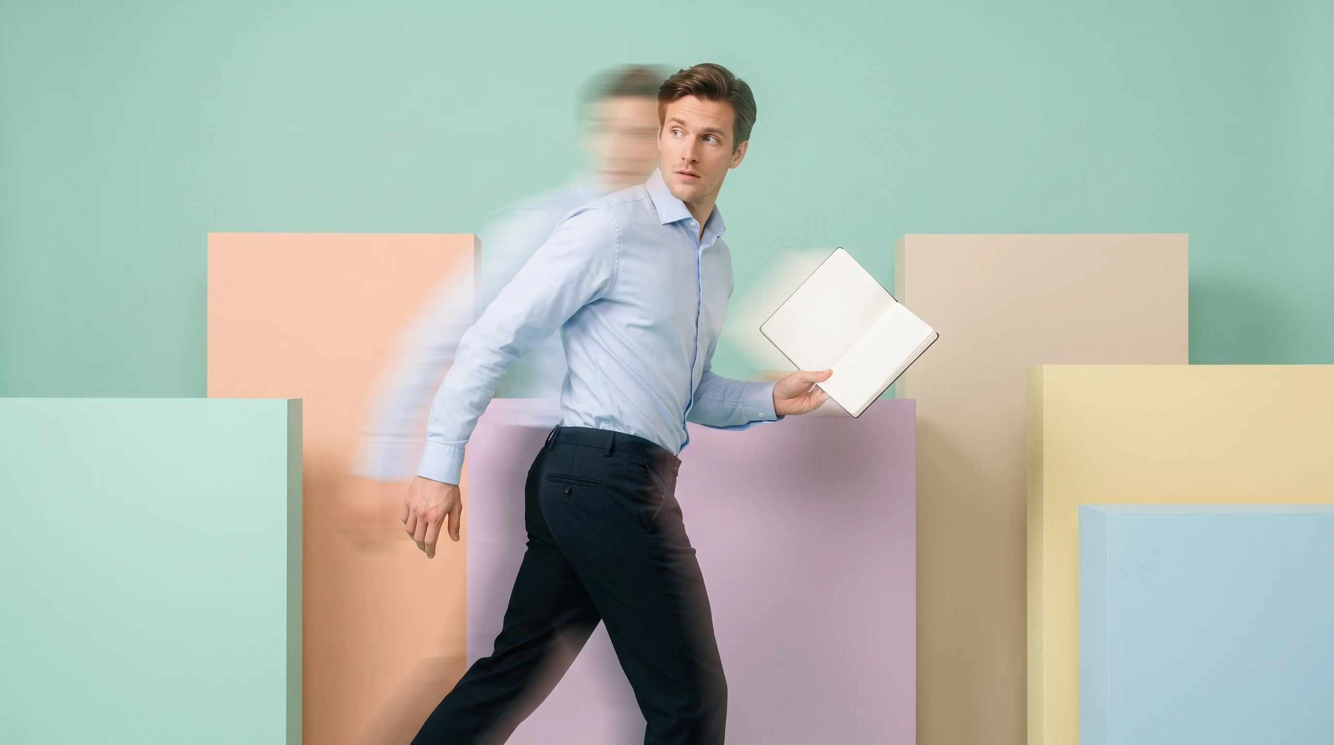 Man mid-step through a mint-backdrop studio, open notebook in hand, glancing back over one shoulder, business-casual shirt, profile framing