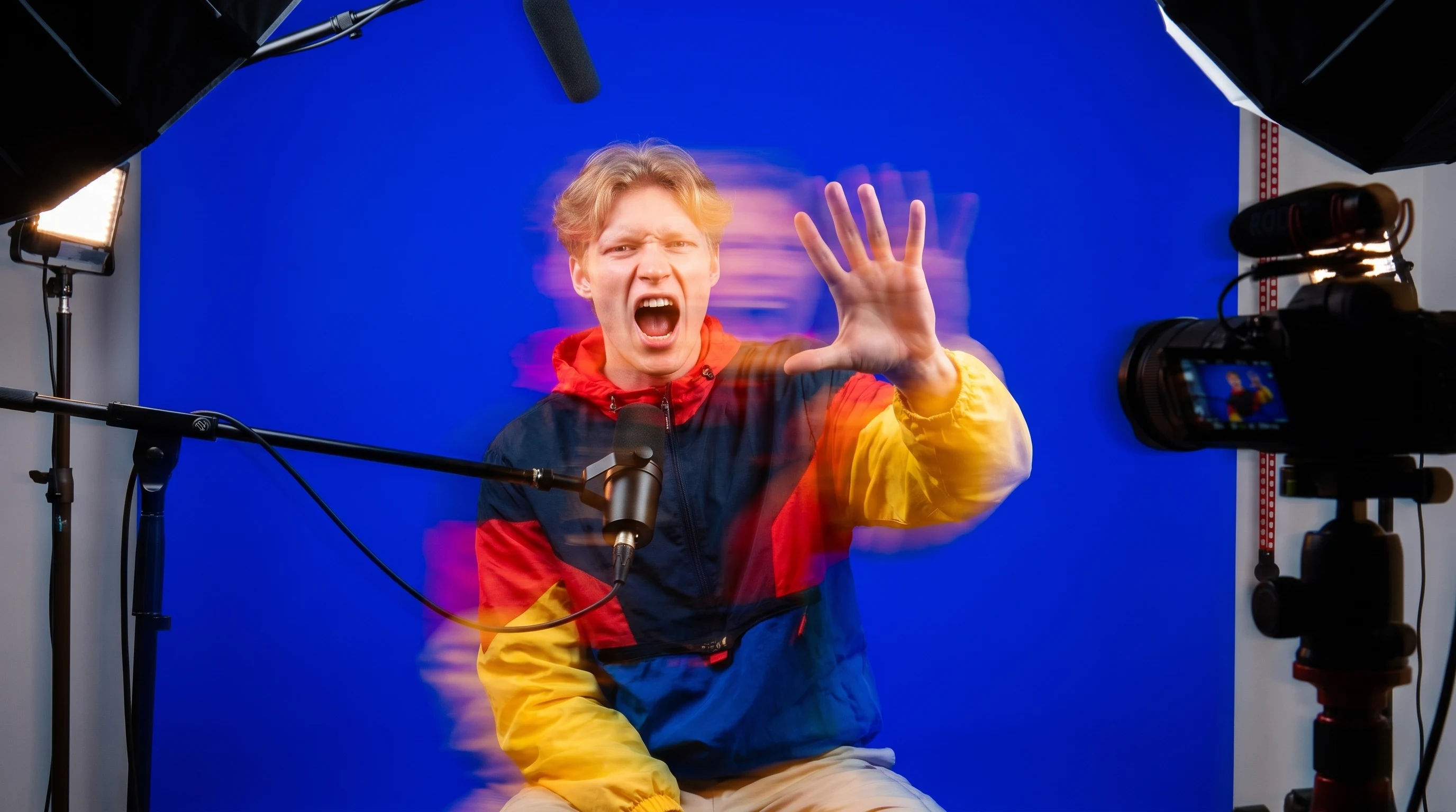 A person mid-gesture with one raised hand facing camera in a compact creator studio, mouth open mid-sentence as if delivering a punchy hook line