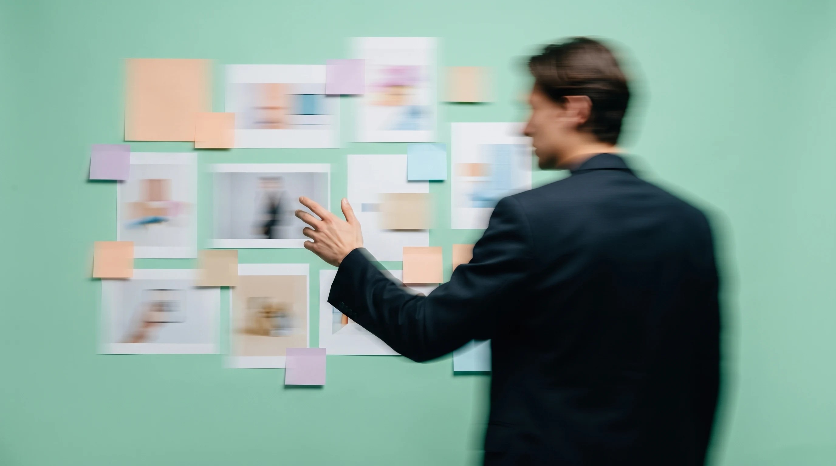 A person mid-turn in a mint-backdrop studio, three-quarter framing, gesturing toward a wall of unbranded concept prints and coloured sticky notes,