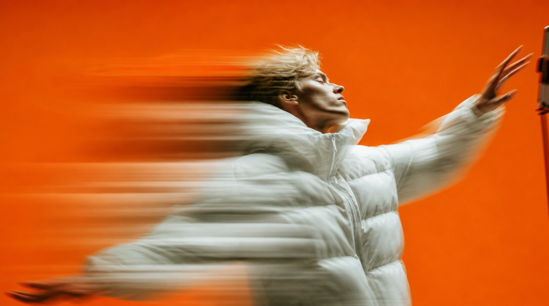 A person mid-turn in a vivid colour-block studio, one arm raised gesturing toward a phone stand just out of frame, shot from slightly below