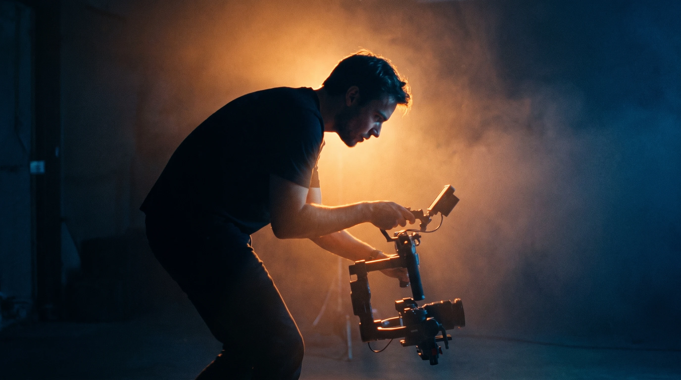A filmmaker mid-step leaning forward to adjust a lightweight camera rig in a stripped-back production space, shot in close profile