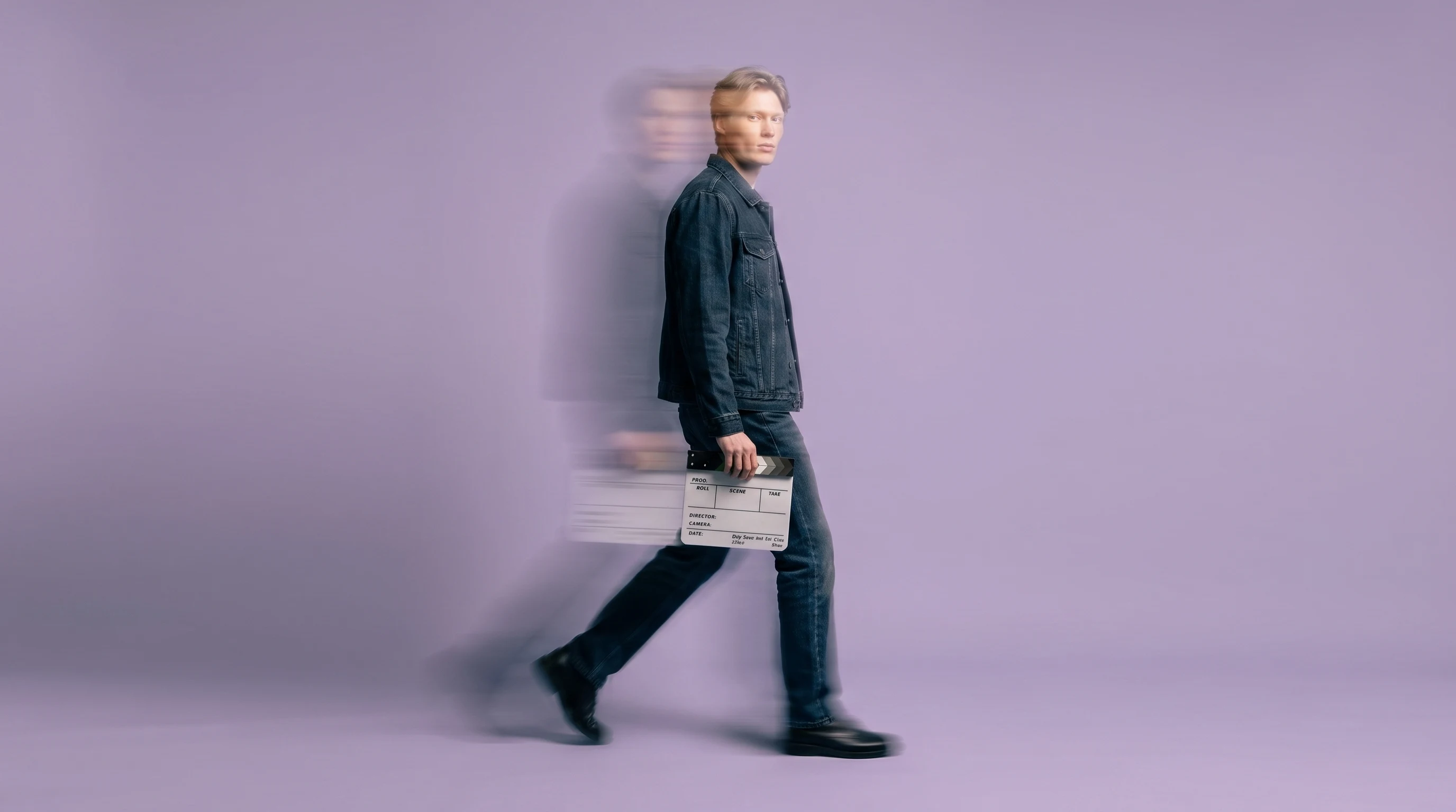 A person mid-step across a lilac-backdrop studio in a dark denim outfit, holding a small clapperboard-shaped prop at hip height, head angled slightly