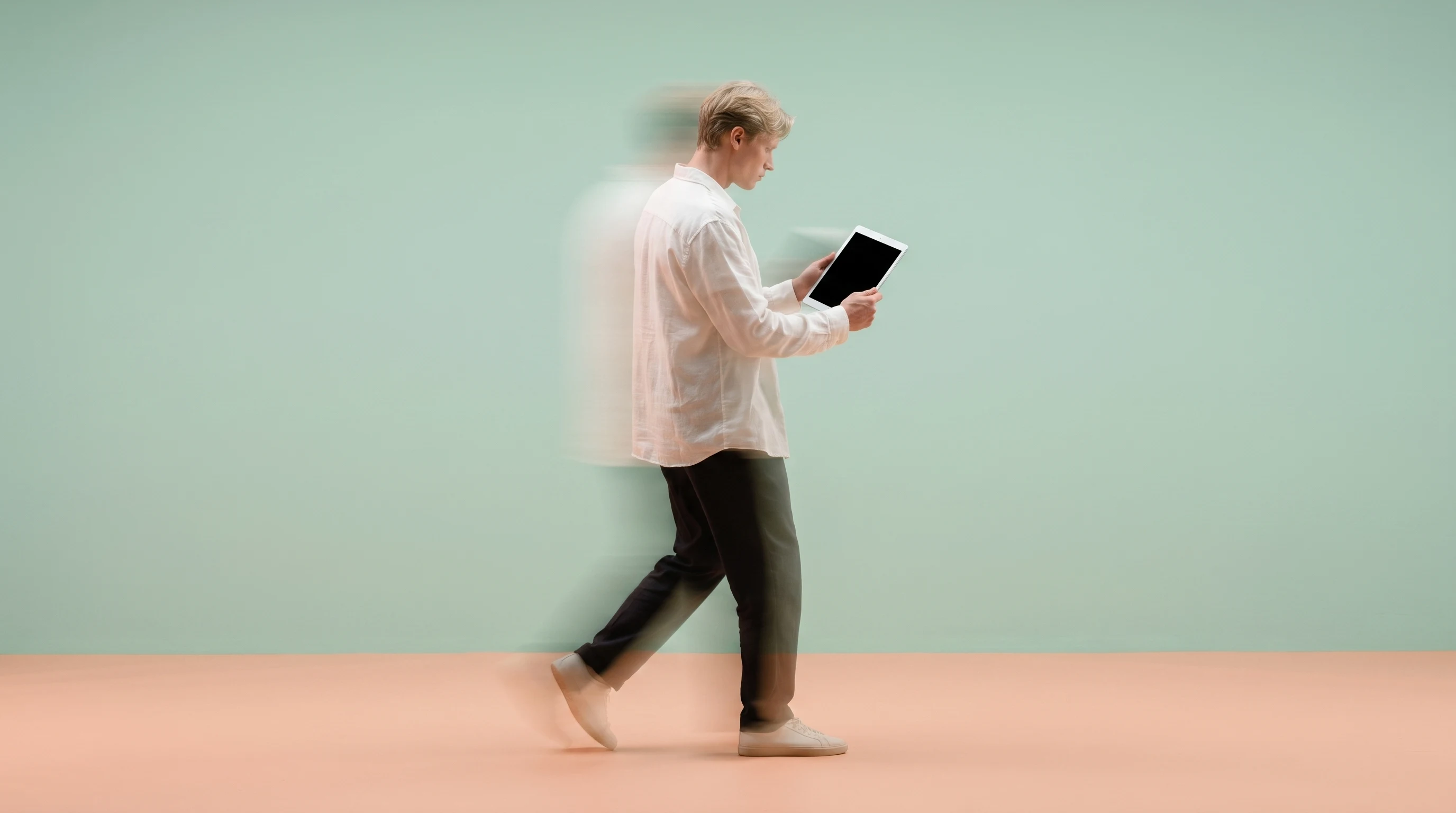 A person mid-step in a peach studio, holding a blank-backed tablet at chest height, glancing down as if reviewing footage frame-by-frame, outfit a