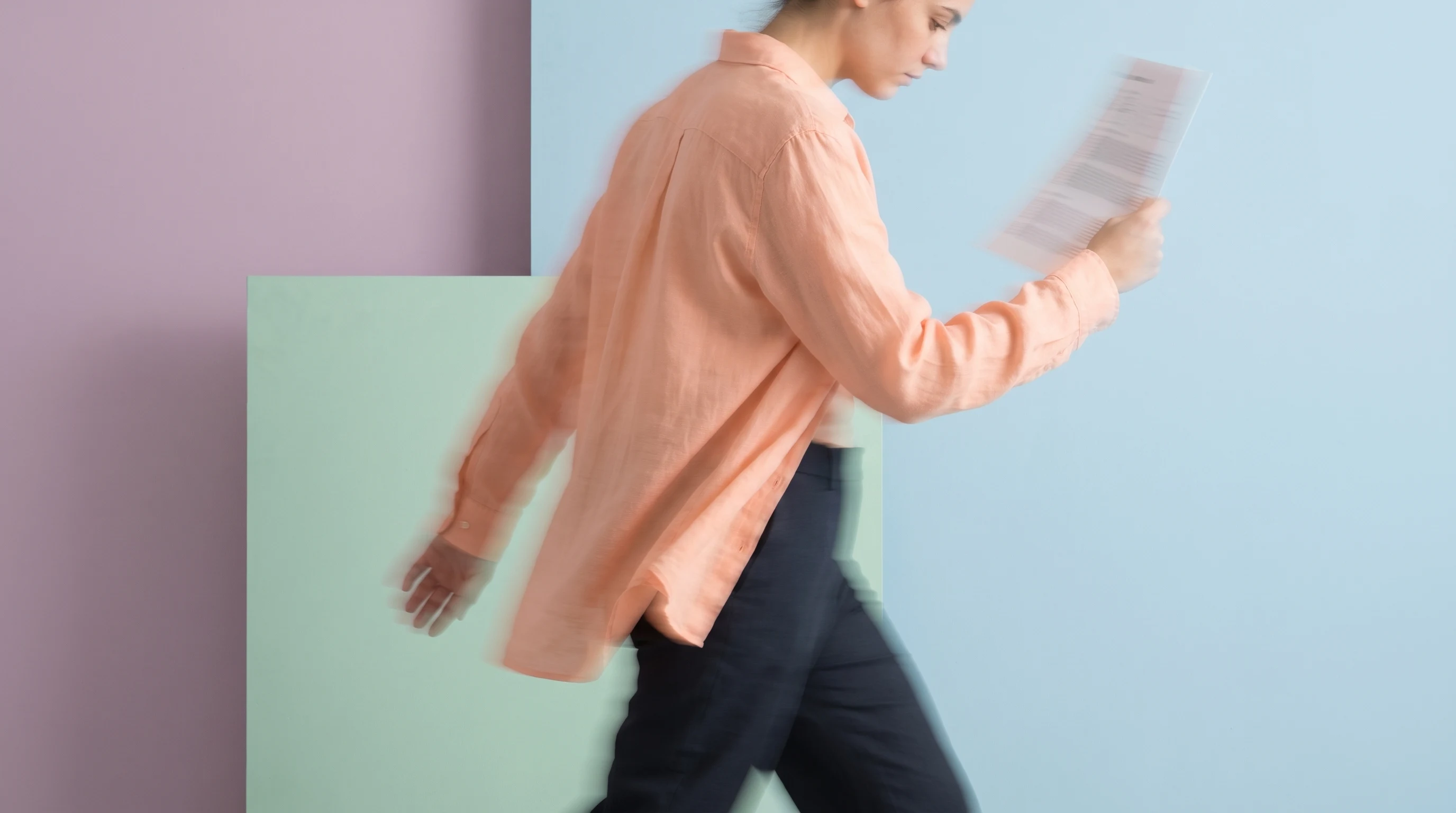 A person in a peach linen shirt mid-step across a lilac-backdrop studio, head slightly bowed reviewing a document held in one raised hand, other arm