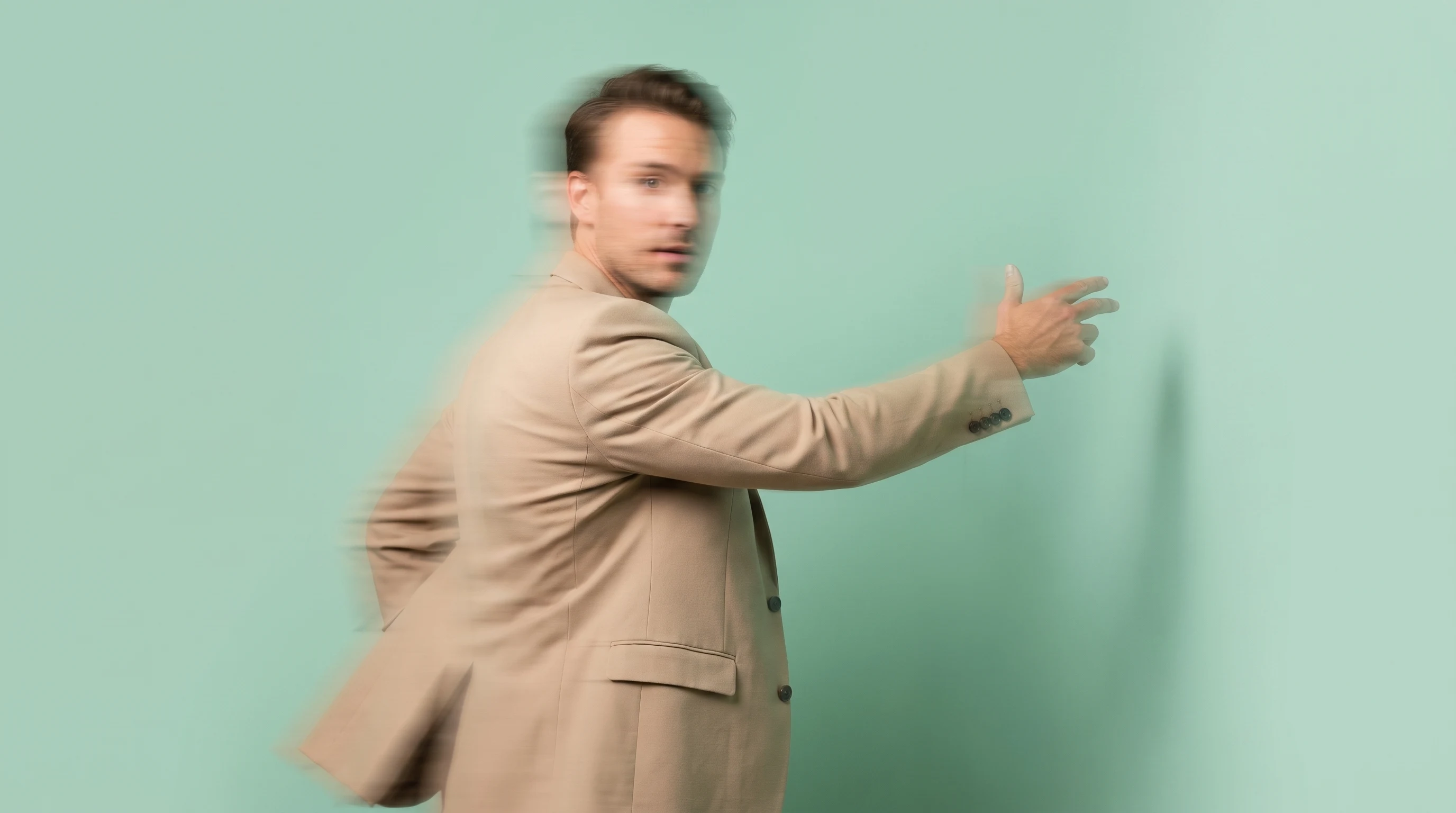 A person in a tailored sand-coloured blazer mid-turn toward camera, one arm extended gesturing at an unseen wall, in a clean mint-backdrop studio