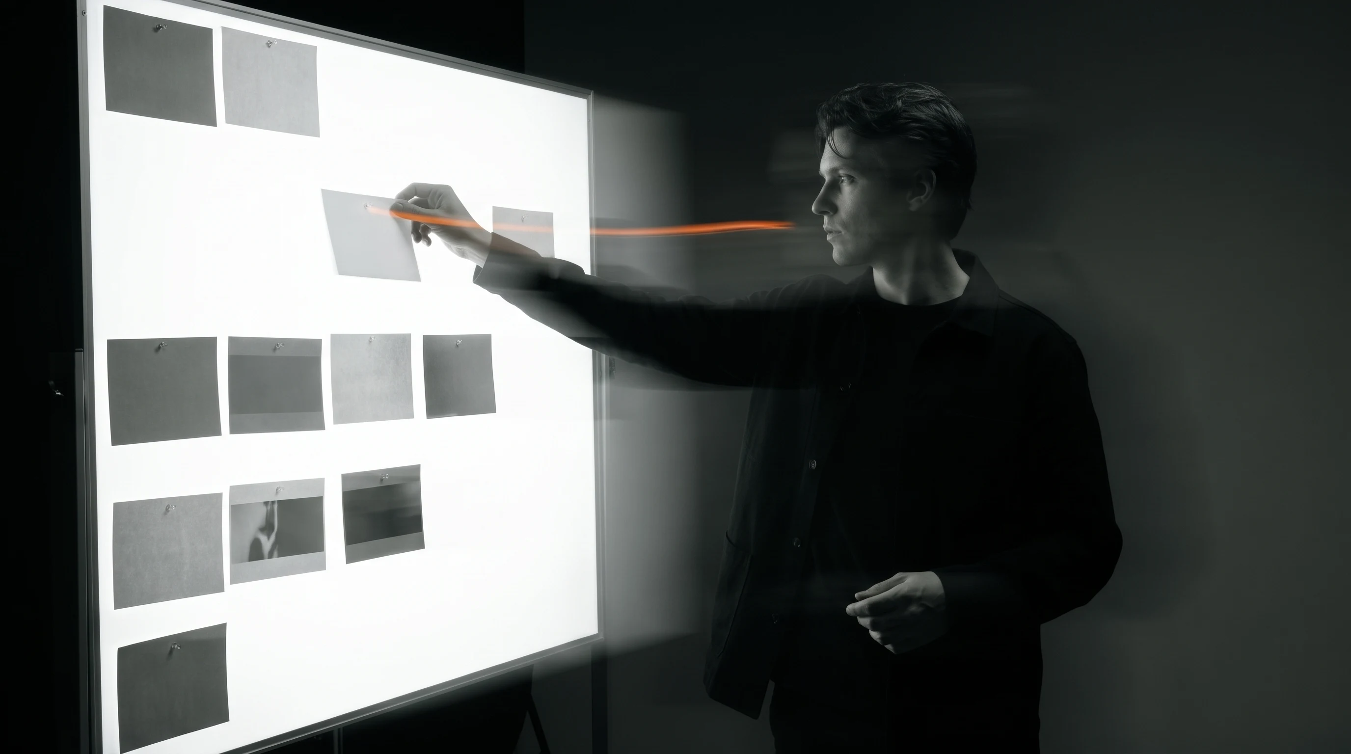 A filmmaker mid-reach in a studio, three-quarter view, pinning an unbranded shot card onto a large backlit sequencing board