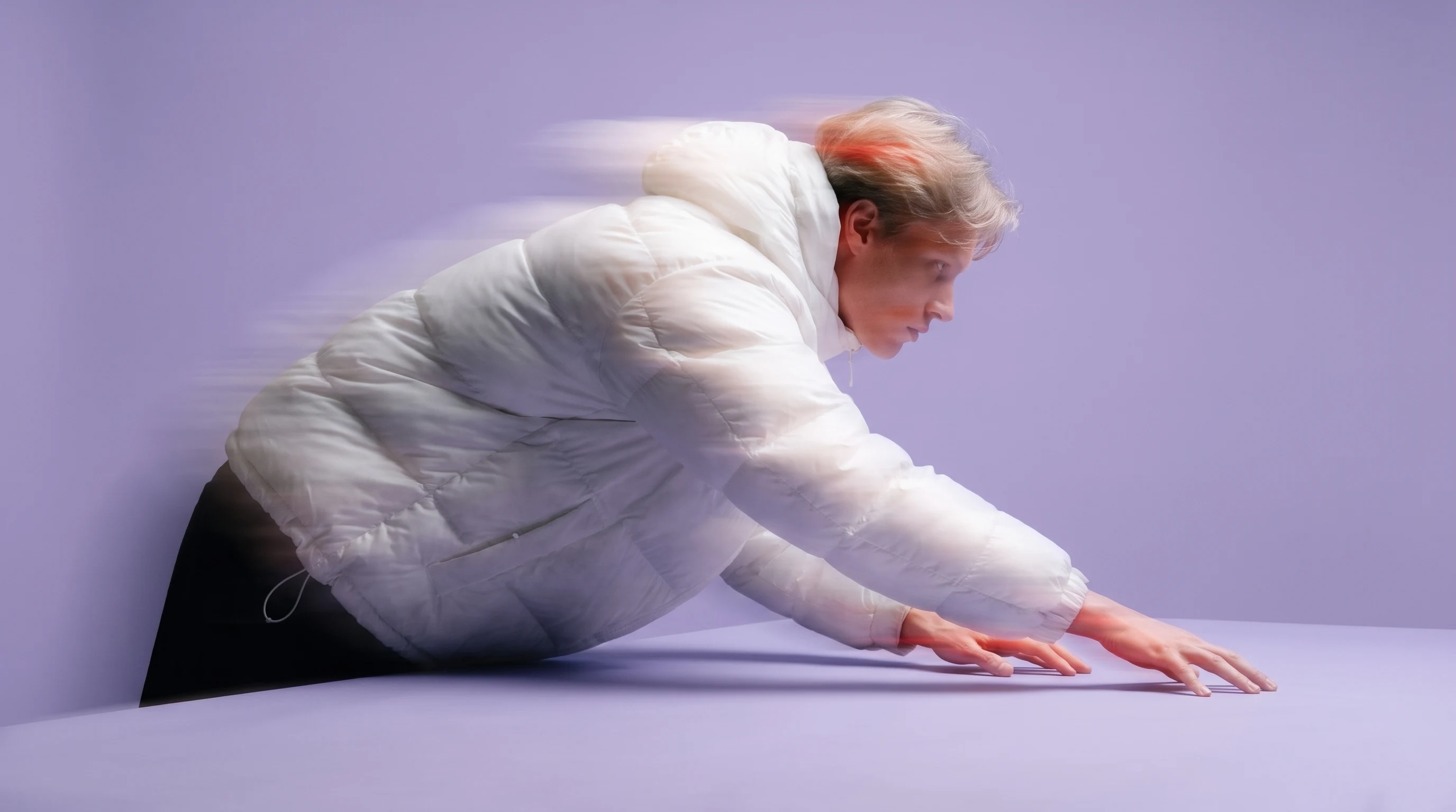 A person mid-reach in a lilac studio, leaning forward over a wide desk, fingers splayed as if curating and arranging a spread of unseen compositions,