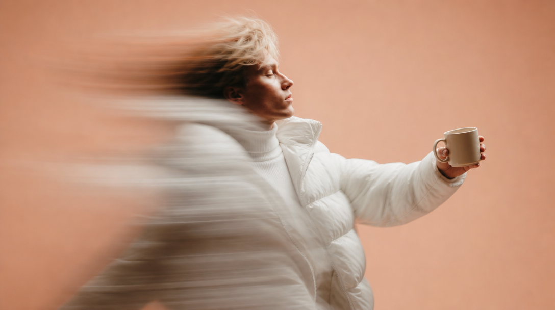 a person mid-turn in a peach-backdrop studio, holding a small unbranded ceramic mug at arm's length toward a softbox just outside the frame, jacket
