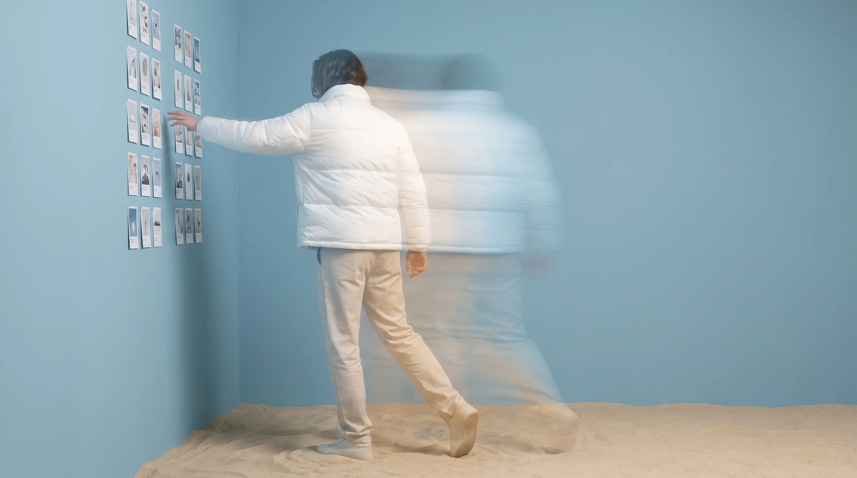 A person in a sand-backdrop studio mid-step forward, three-quarter framing from behind, reaching toward a wall of small unbranded printed cards