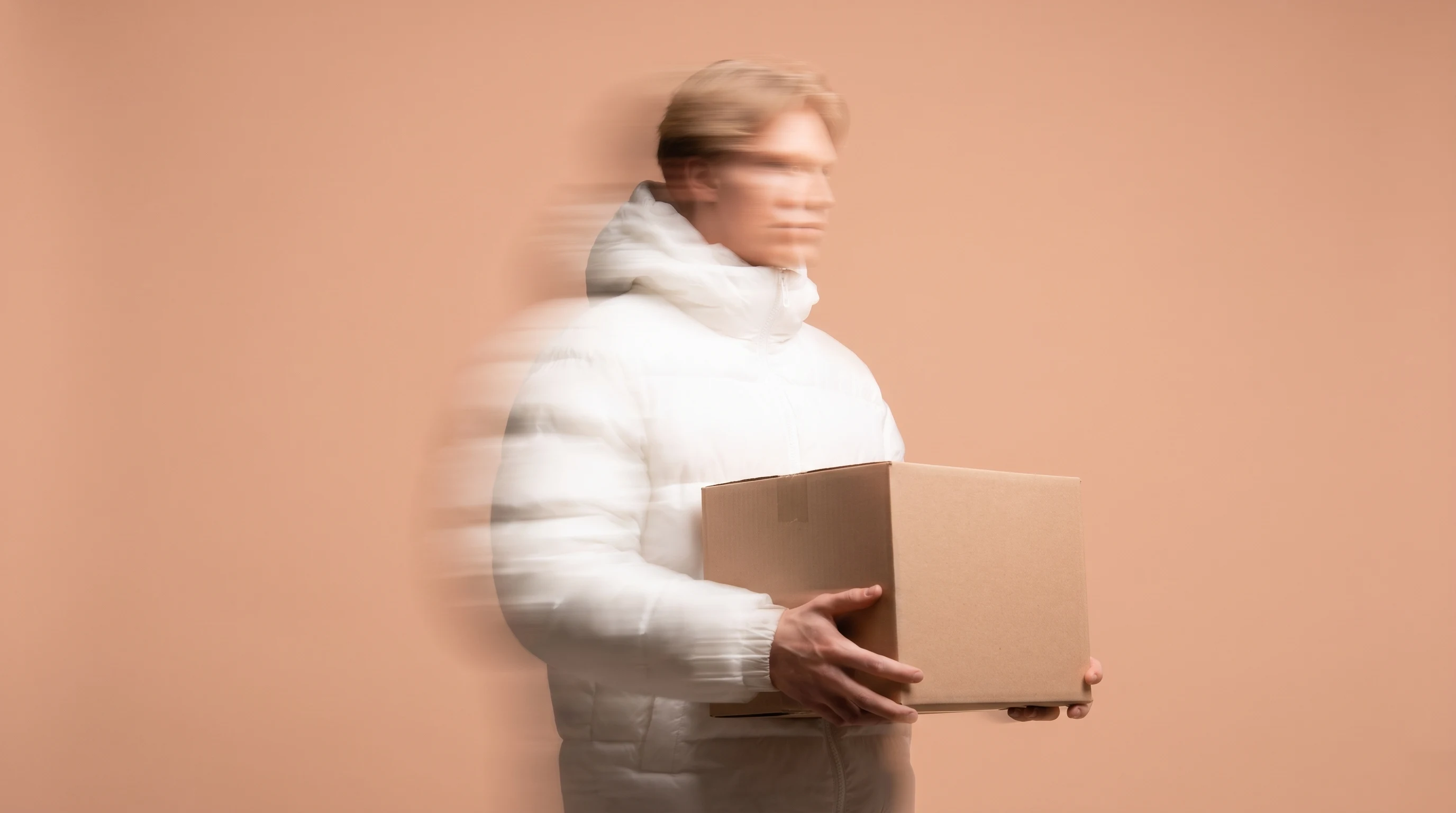 A person mid-turn in a peach-backdrop studio, holding a blank unbranded box at waist height, three-quarter framing from the front