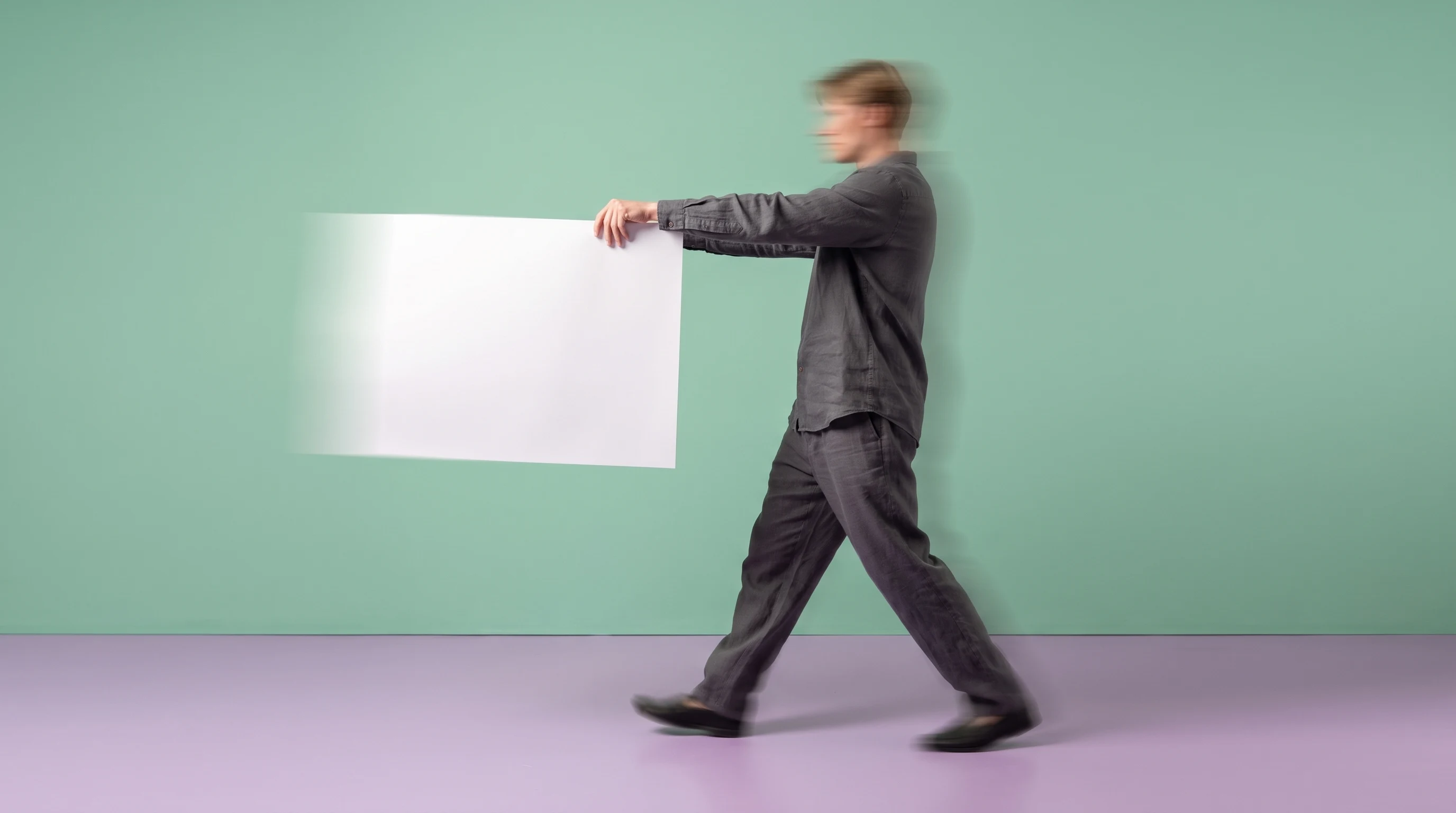 A person mid-step across a lilac studio floor, holding a large unbranded print at arm's length toward camera, profile framing, wearing a loose