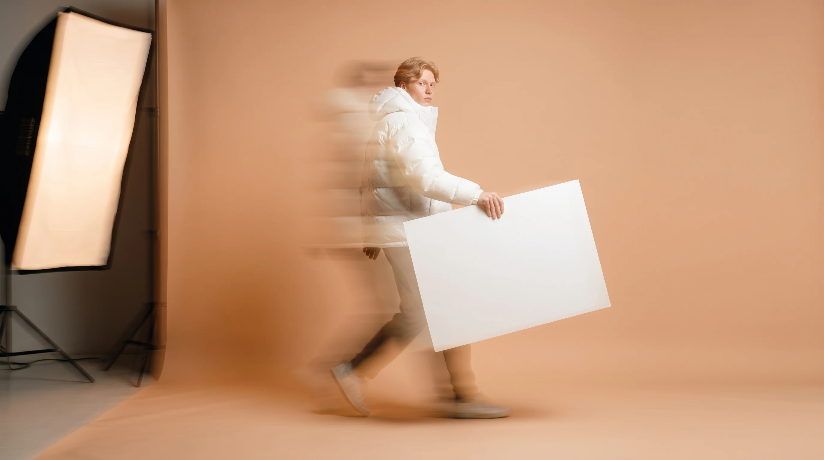 A person mid-step across a peach-backdrop studio, carrying a large blank white portfolio board at arm's length, glancing back over one shoulder